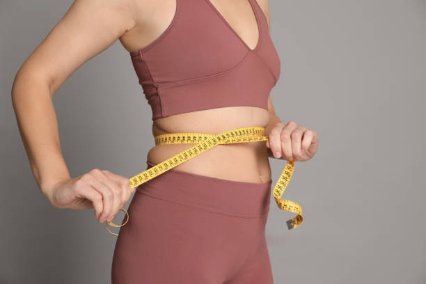 woman measuring waist with tape on grey background, closeup